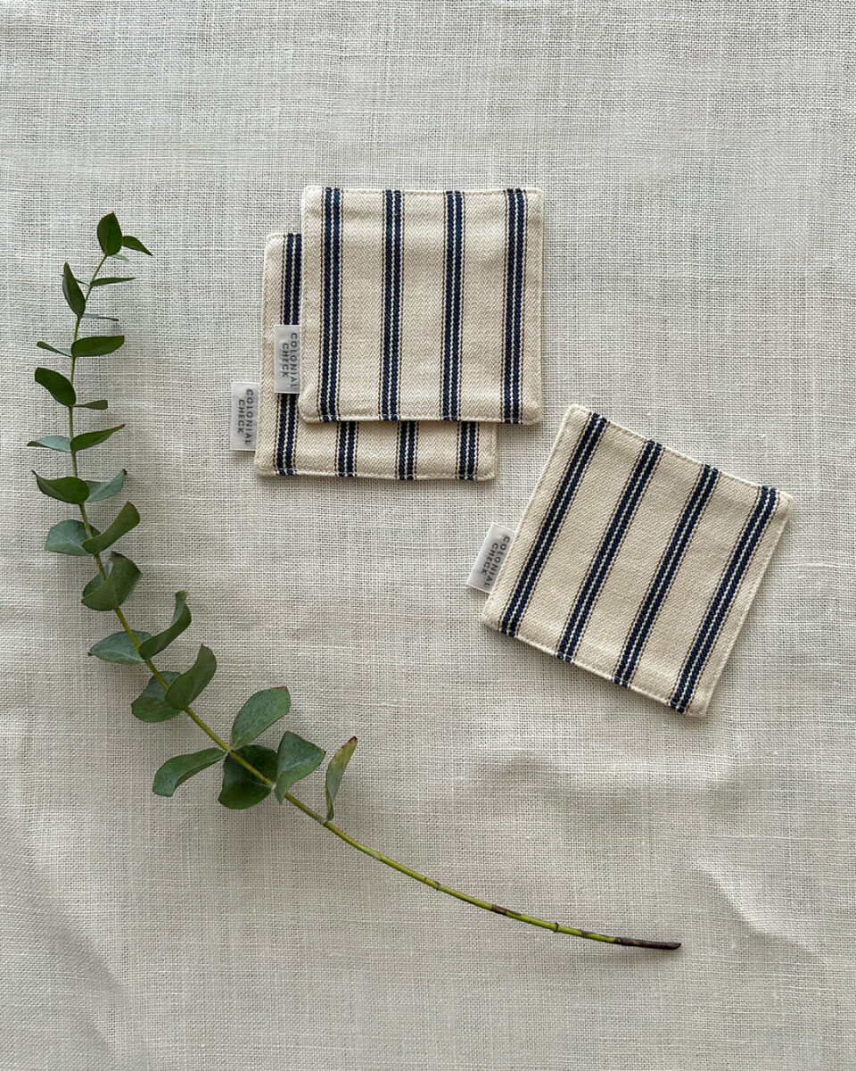 Three beige napkins with blue stripes, folded on a light linen background, accompanied by a eucalyptus stem for a clean, natural look.