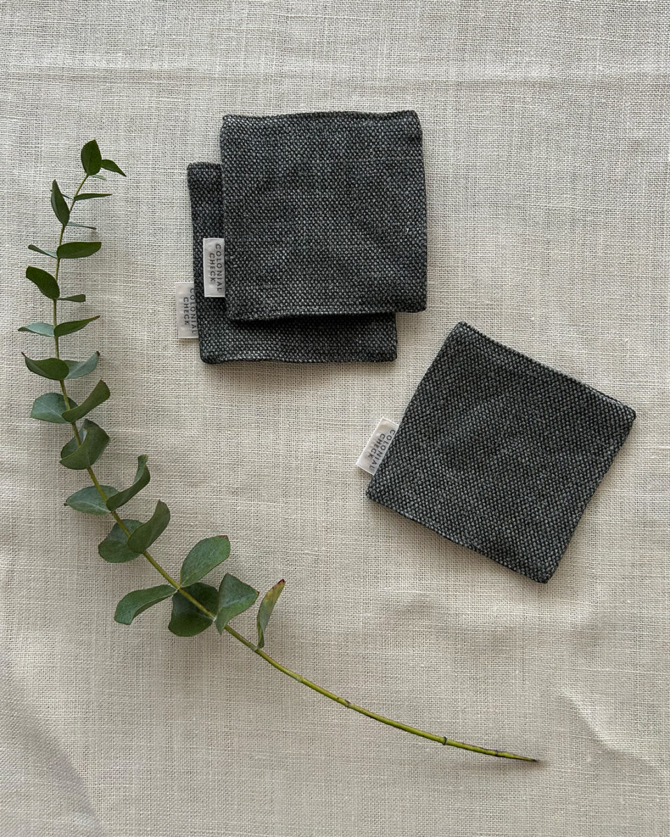 Three charcoal gray fabric swatches with white tagged labels on a light linen background, accompanied by a eucalyptus sprig across the fabric.