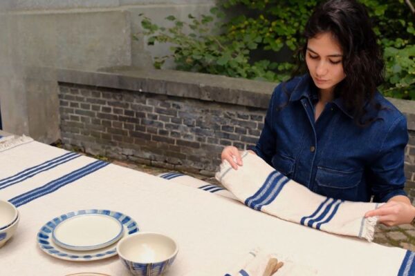 Woman in a denim shirt outdoors folds a white cloth with blue stripes over a long table set with blue-and-white dishes and bowls on a stone terrace.