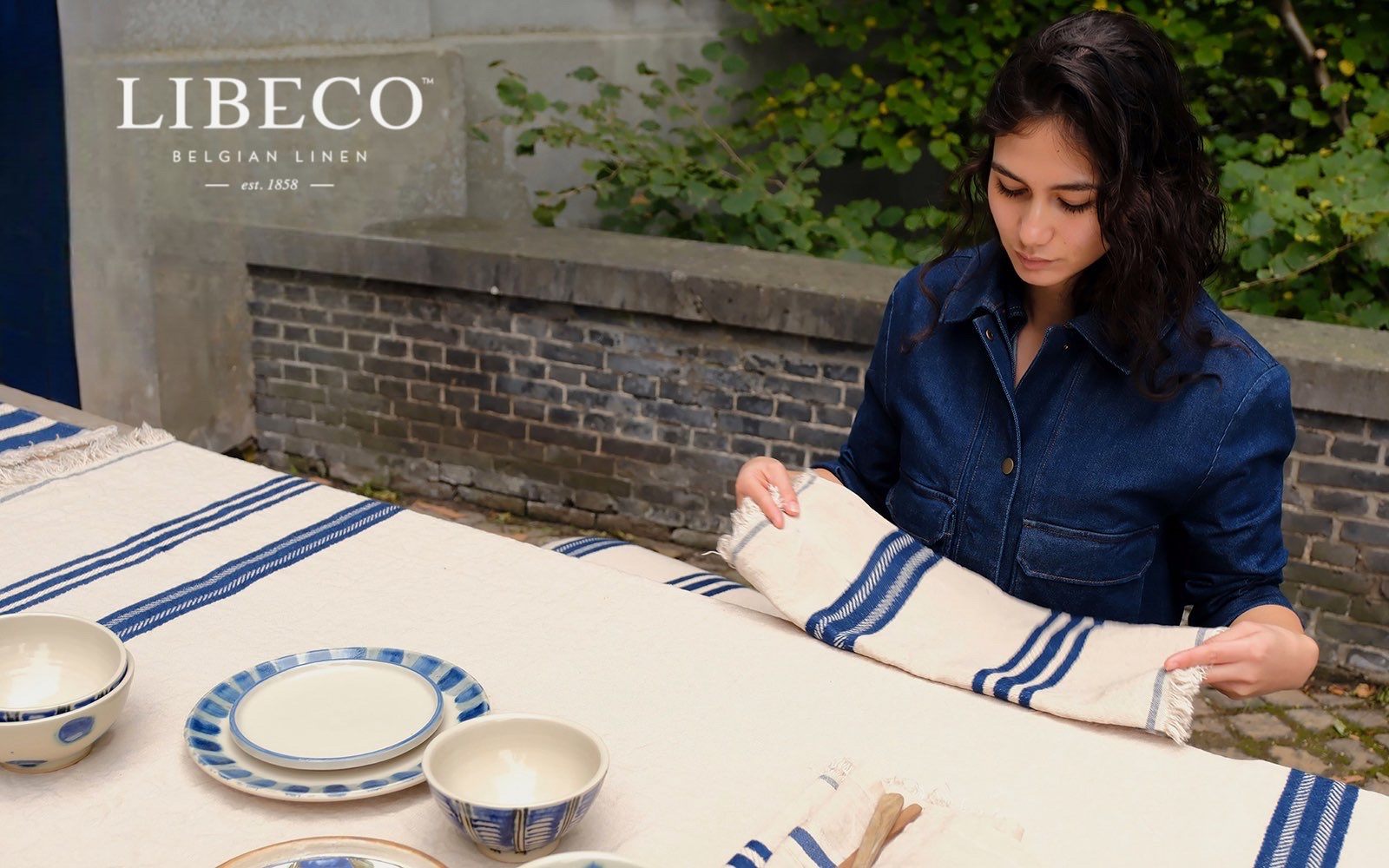 Woman in a denim shirt folds a cream linen towel with blue stripes on an outdoor table with ceramic bowls nearby.