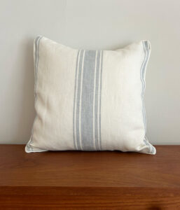 White decorative pillow with blue vertical stripes resting on a wooden surface against a pale wall, front view.