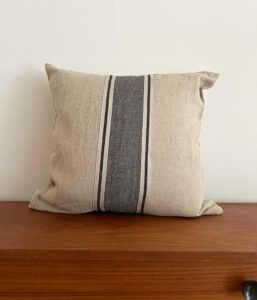 Beige fabric throw pillow with a dark gray striped center resting on a wooden surface against a pale wall.