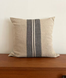 Beige cushion with dark vertical stripes resting on a wooden surface against a pale wall.