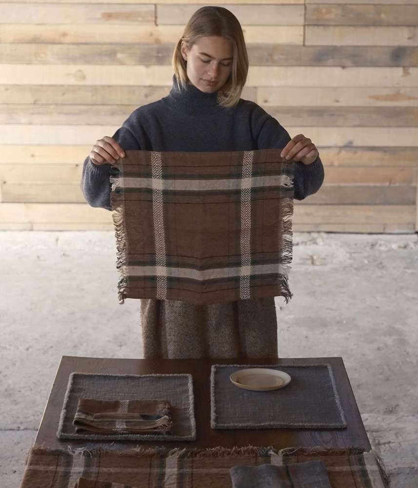 Woman in a blue sweater holds up a brown plaid scarf with fringed edges in a workshop setting.