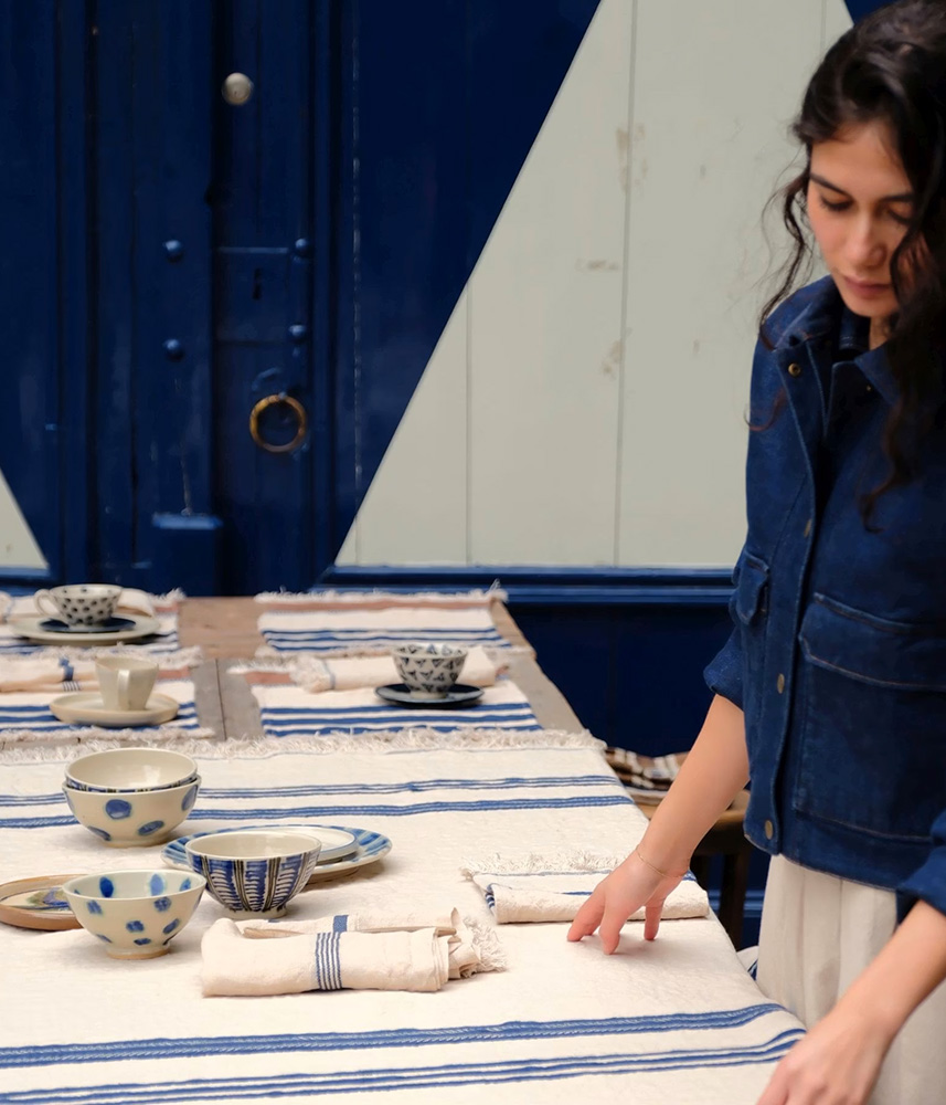 Person in a denim jacket arranges blue-and-white ceramic bowls and napkins on a striped tablecloth, setting an outdoor dining table.