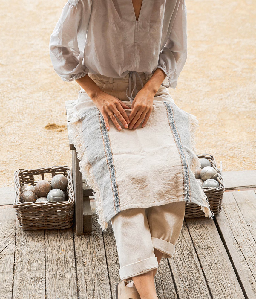 Woman in light linen clothing sits on a wooden deck with baskets of pétanque boules beside her.