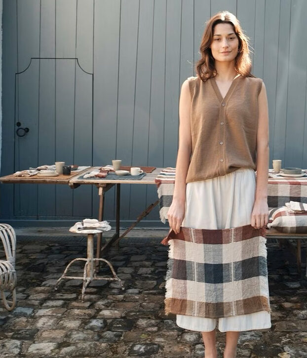 Woman standing in a sunny courtyard next to a table set with dishes, wearing a sleeveless tan top and white skirt. A wicker chair sits nearby against a gray wall.