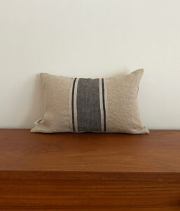 Beige rectangular decorative pillow with a dark central stripe on a wooden bench against a pale wall.