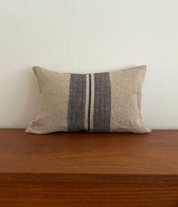 Beige rectangular throw pillow with two dark blue vertical stripes on a wooden surface against a pale wall.