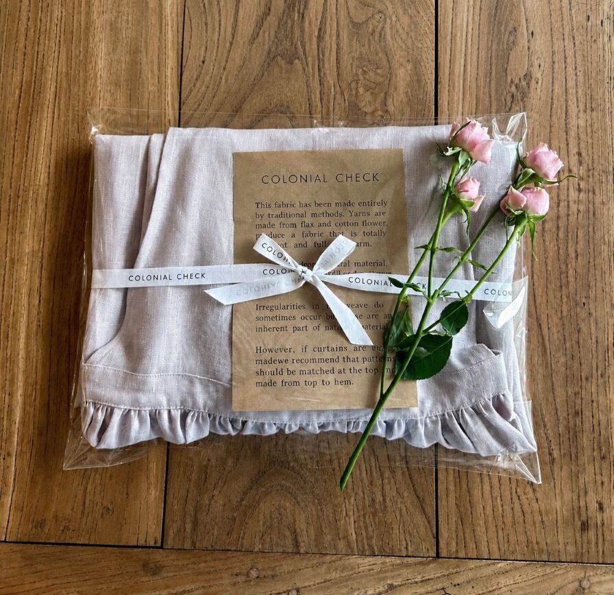 Soft pink fabric folded inside a clear garment bag, tied with a white ribbon reading 'Colonial Check' on a wooden table, with pink roses laid on top.