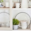 White shelf displaying dishes and glassware with small potted plants, bowls, and a pitcher in a bright kitchen.