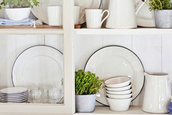 White shelf displaying dishes and glassware with small potted plants, bowls, and a pitcher in a bright kitchen.