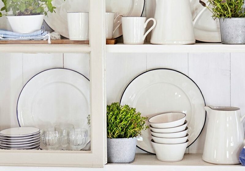 White shelf displaying dishes and glassware with small potted plants, bowls, and a pitcher in a bright kitchen.