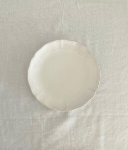 Top-down view of a plain white ceramic plate with a slightly scalloped edge on a light fabric surface.
