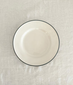 Top-down view of a plain white ceramic plate with a dark rim on a light fabric tablecloth