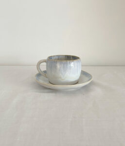 Small ceramic cup with pale blue glaze on a matching saucer, set on a white tablecloth against a pale wall.