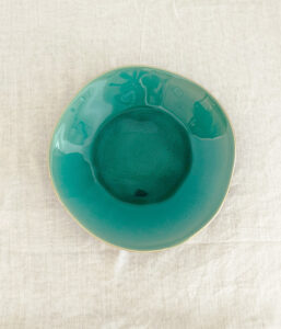 Round teal ceramic bowl with a shallow inner well, photographed from above on a light beige cloth.