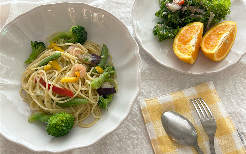 Plate of spaghetti with shrimp and mixed vegetables (broccoli, peppers) on a white dish; kale salad with orange wedges on the side.