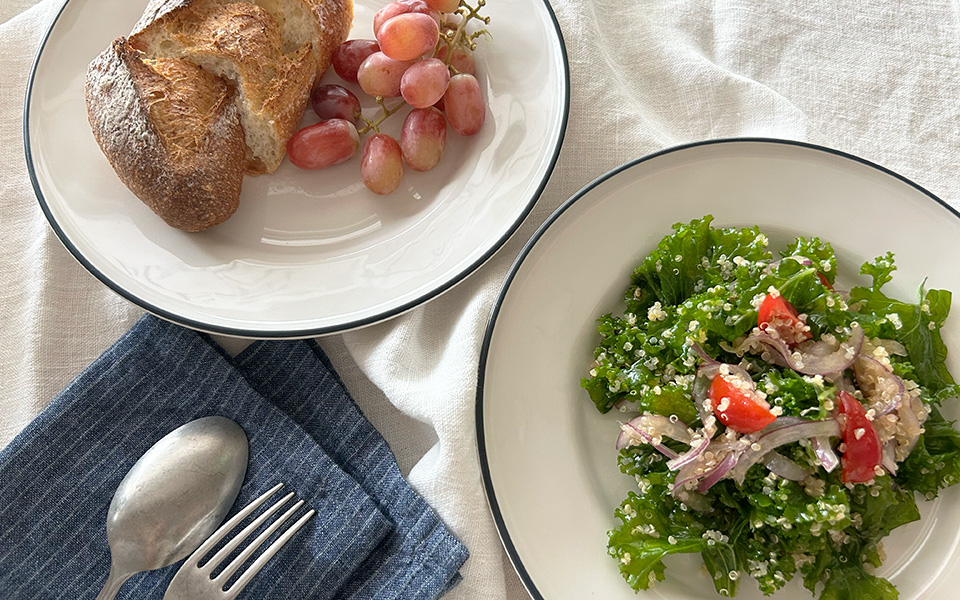 Two white plates hold bread with a bunch of red grapes on one plate and a kale salad with tomatoes, onions, and feta on the other; a spoon and fork rest on a blue napkin nearby.