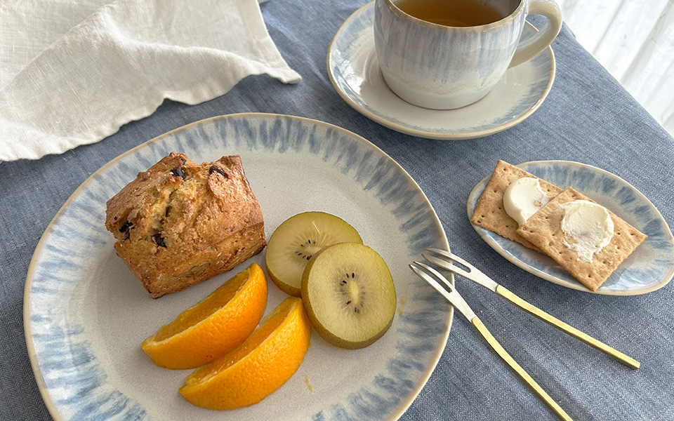 Plate with a muffin, orange wedges, and kiwi slices on a blue-rimmed plate; a cup of tea nearby, and a small plate with crackers and butter.