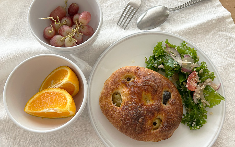 Brunch plate with a toasted bagel and a side of kale salad on a white plate, with orange wedges and a bowl of grapes nearby on a light tablecloth.