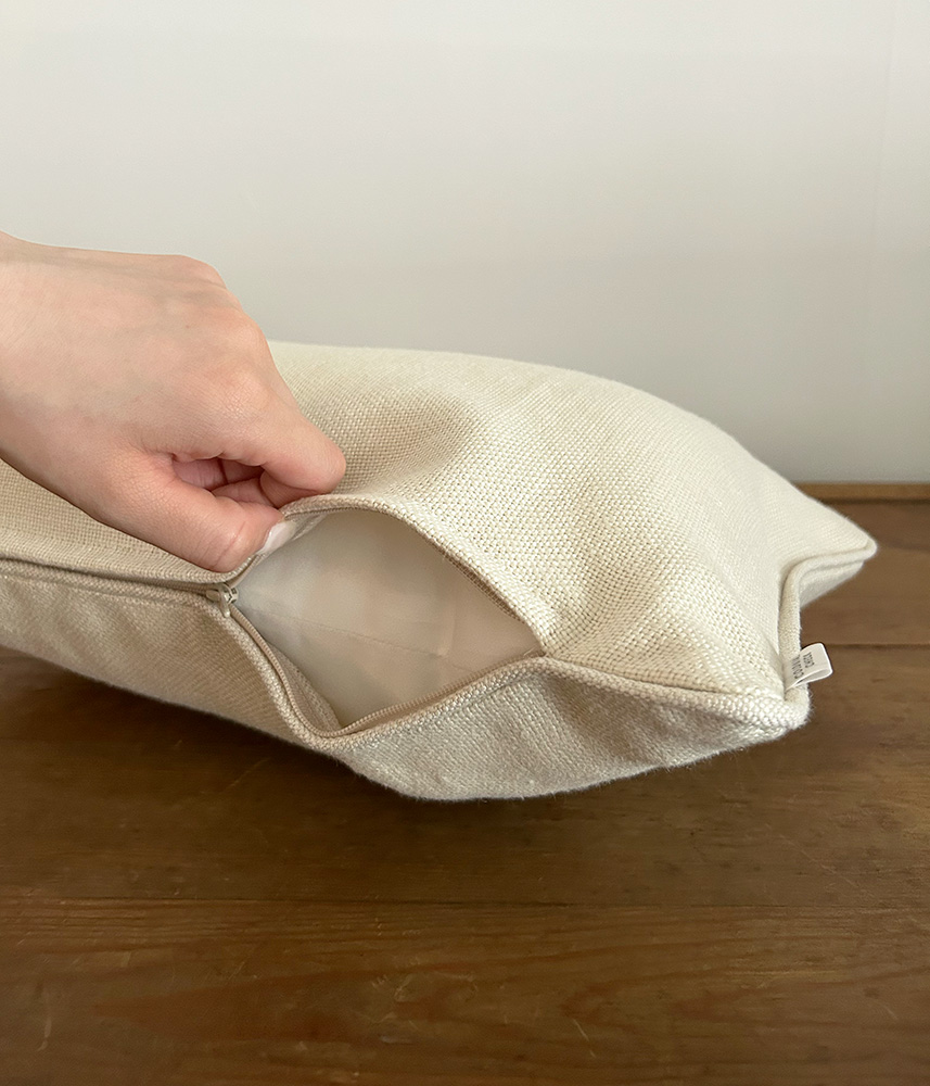 Person opening a beige fabric pillow cover by one corner zipper to insert or remove stuffing.