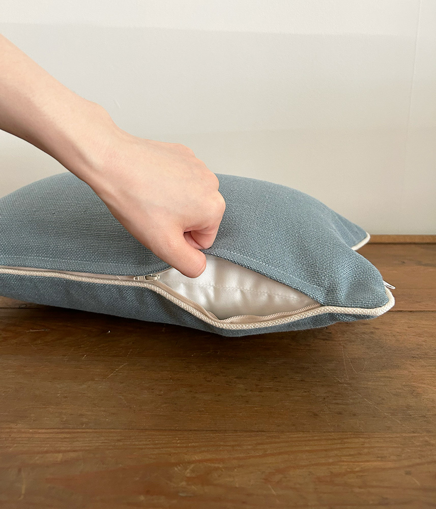 Hand pulling open a blue fabric cushion to reveal the white inner lining and zipper along the edge on a wooden table.