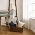 Wooden stool with stacked cushions, a wicker basket of pillows, a ladder with a striped towel, by a sunlit window.